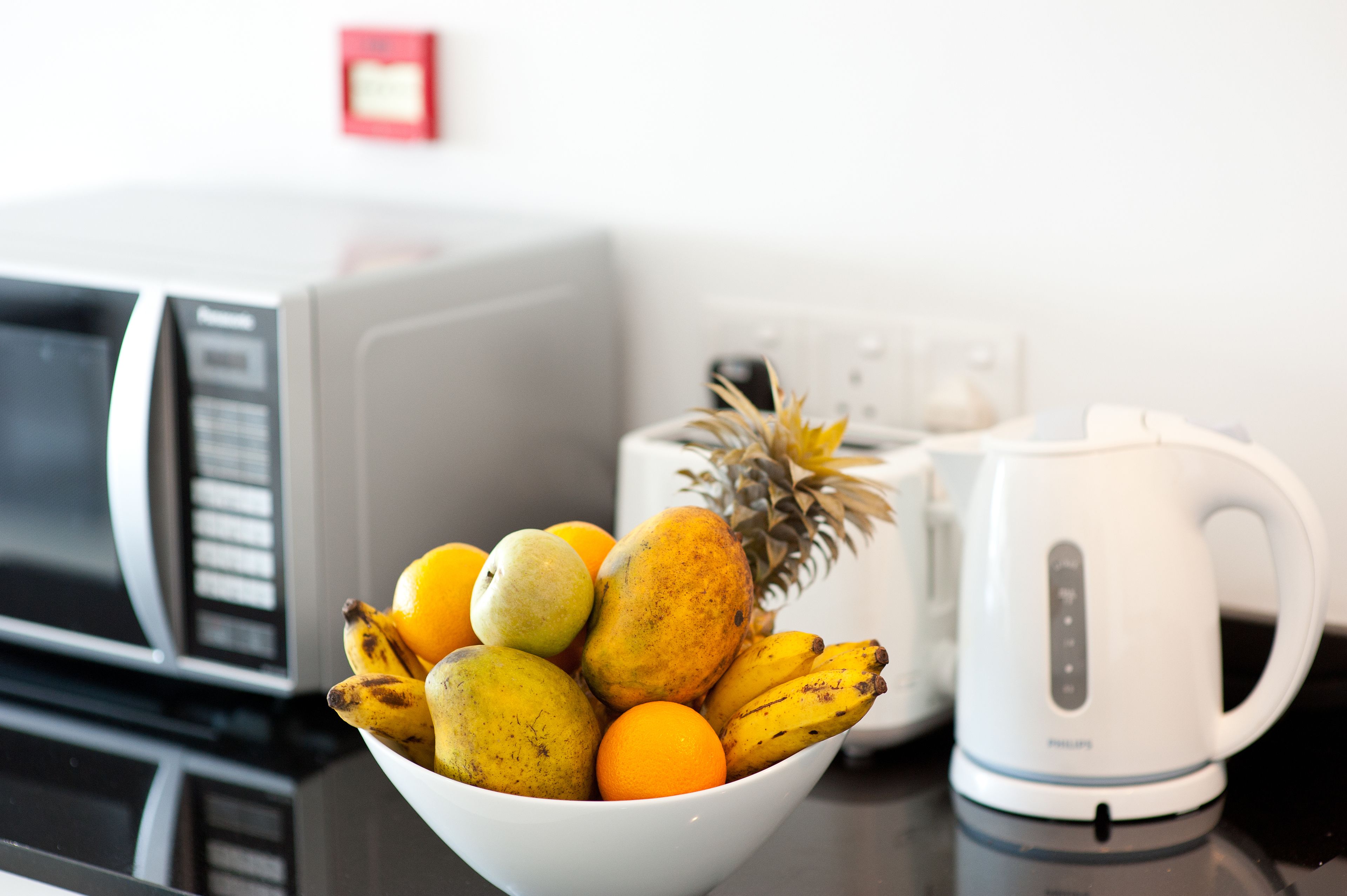 Una encimera de cocina con un bol blanco lleno de fruta, una cafetera blanca y un microondas.