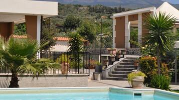 Zona de piscina con vistas a una cadena montañosa, una escalera que lleva a un edificio y diversas plantas y muebles de exterior.