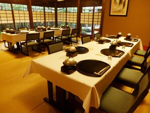 Restaurante com várias mesas preparadas para refeições, com decoração tradicional japonesa e vista para o verde através de grandes janelas.
