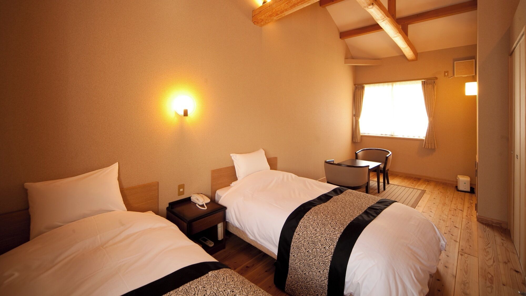 A hotel room with two beds, a small table, a chair, and a window with curtains.