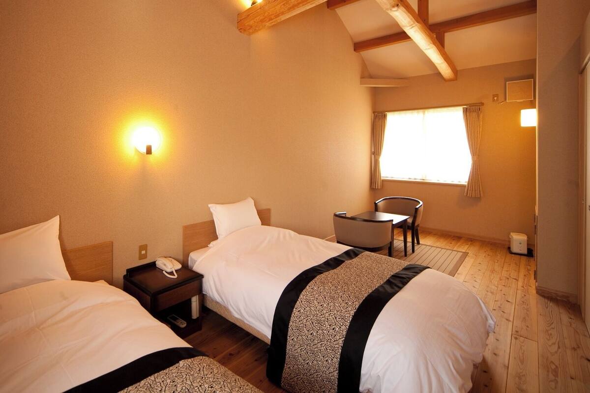 Habitación de hotel con dos camas, una mesita, una silla y un ventanal con cortinas.