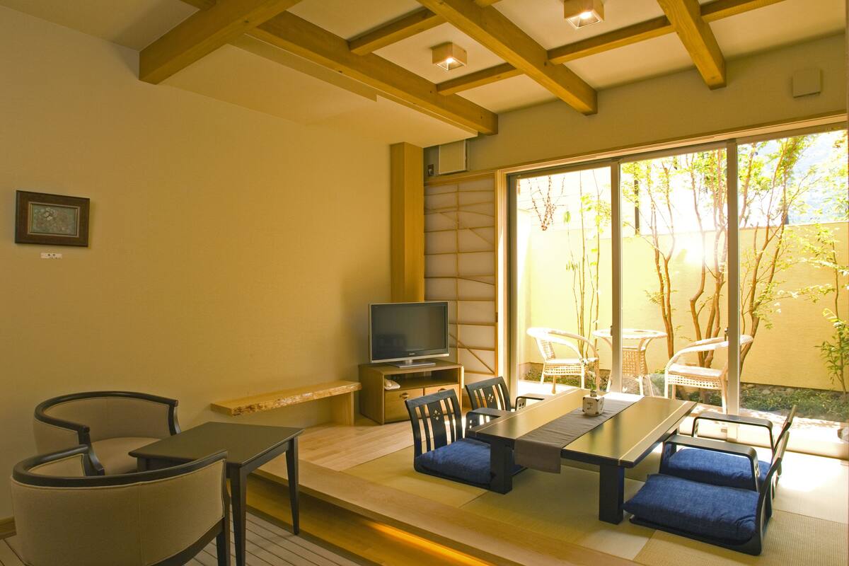 Comedor con una mesa larga, sillas, televisor y puerta corrediza de vidrio que da a un espacio exterior con sillas y plantas.