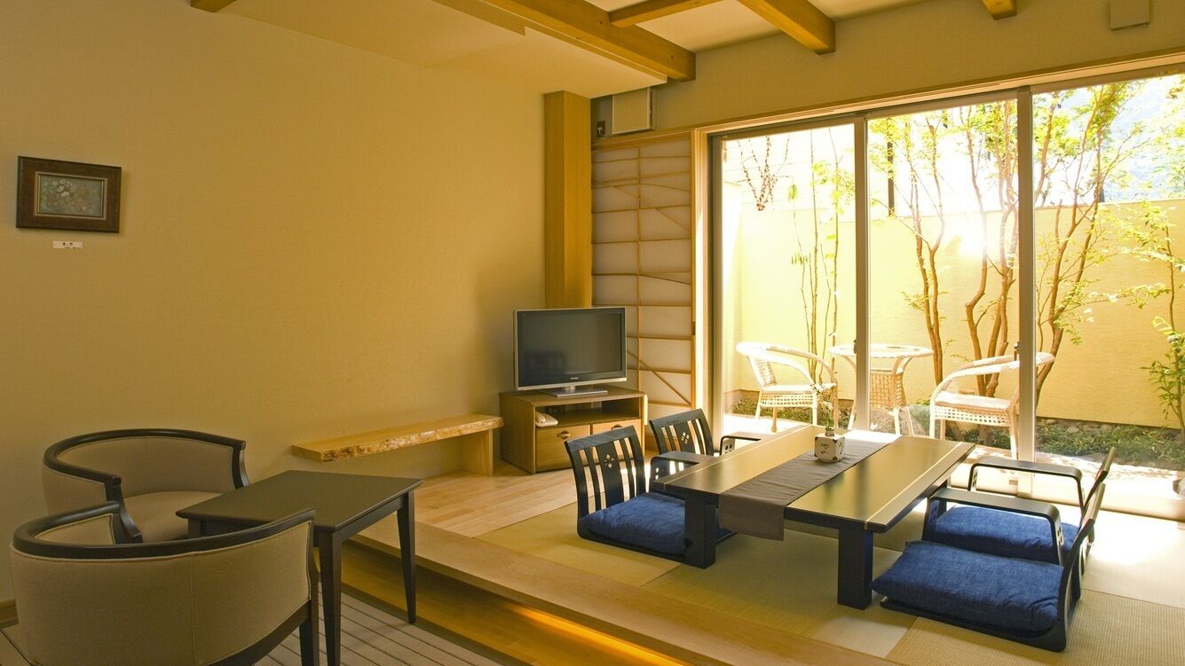 A room with a tatami mat floor, a low table with cushions, a television on a stand, and sliding glass doors leading to an outdoor area with chairs and plants.