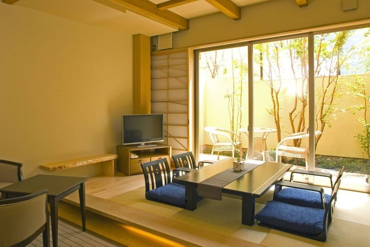 Una habitación con suelo de tatami, una mesa baja con cojines, un televisor sobre un mueble y puertas correderas de cristal que dan a un espacio exterior con sillas y plantas.
