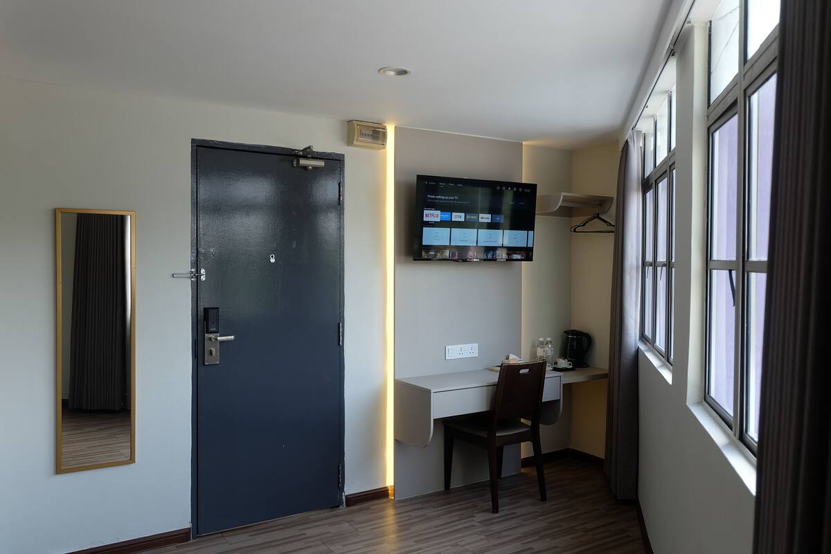 Une chambre d’hôtel avec une porte sombre, un bureau avec une chaise, un téléviseur fixé au mur et une fenêtre avec des rideaux.