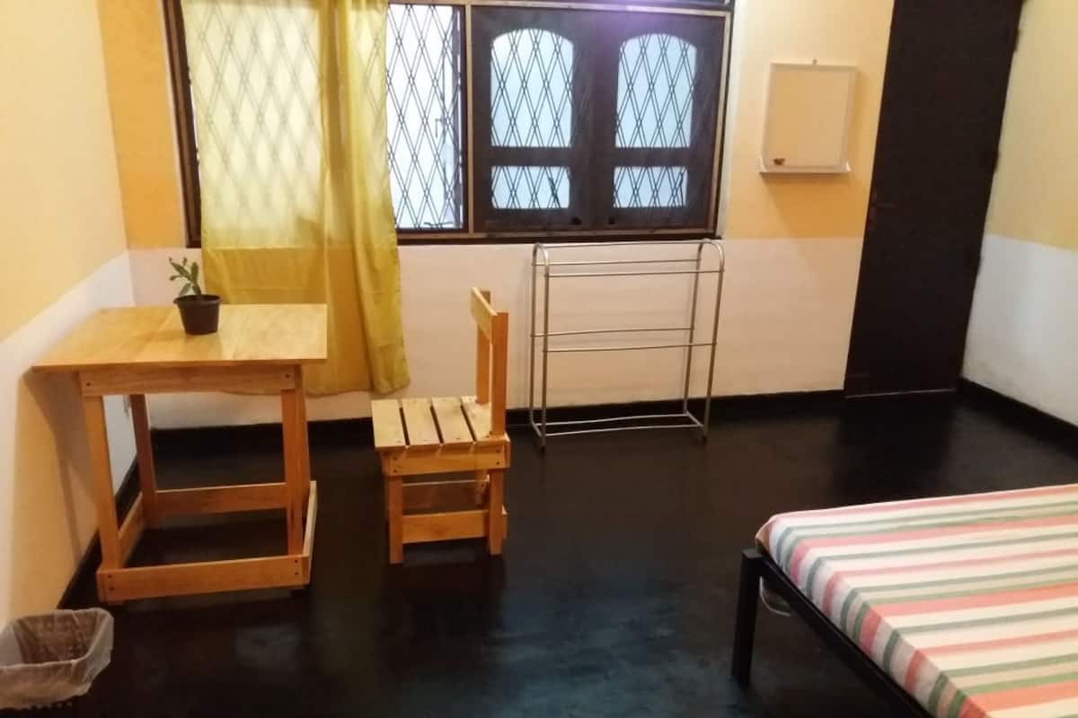 A small, simple room with a bed, a wooden chair, a small table, and a window with a curtain.