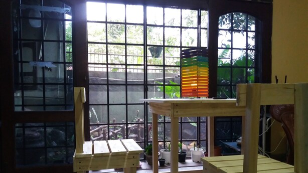 Een kamer met een groot raam, houten stoelen, een kleine tafel en planten in potten.