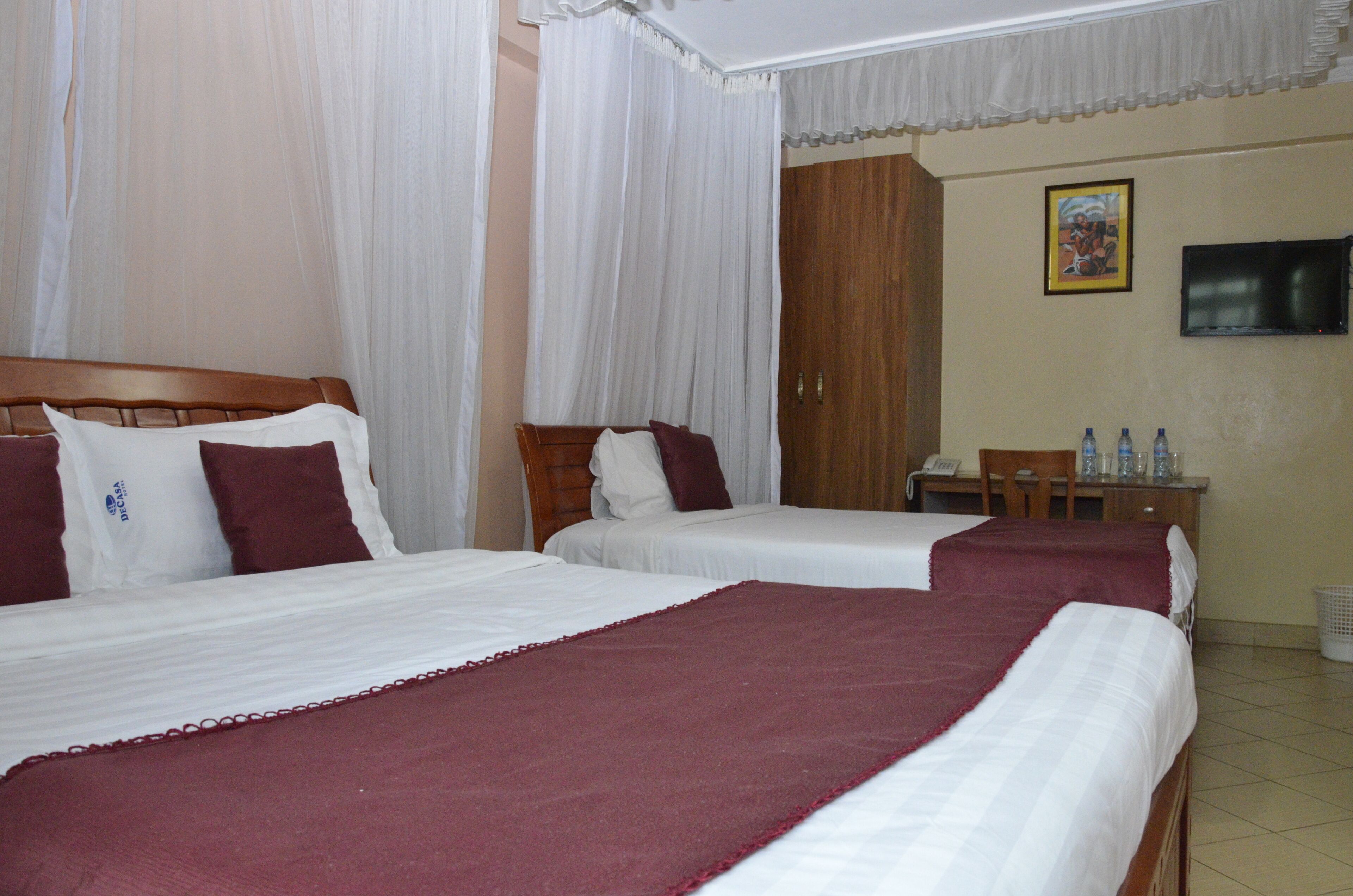 A hotel room with two beds, white bedding, a wooden headboard, a television on the wall, and a small table with water bottles.