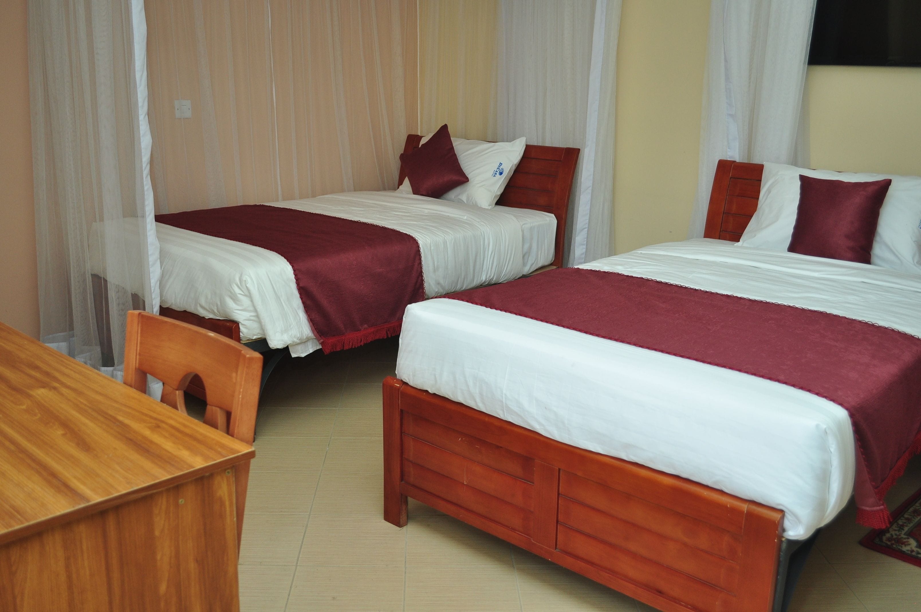 Une chambre d’hôtel avec deux lits en bois, chacun agrémenté de draps blancs et de touches de couleur rouge, un bureau en bois et une chaise.