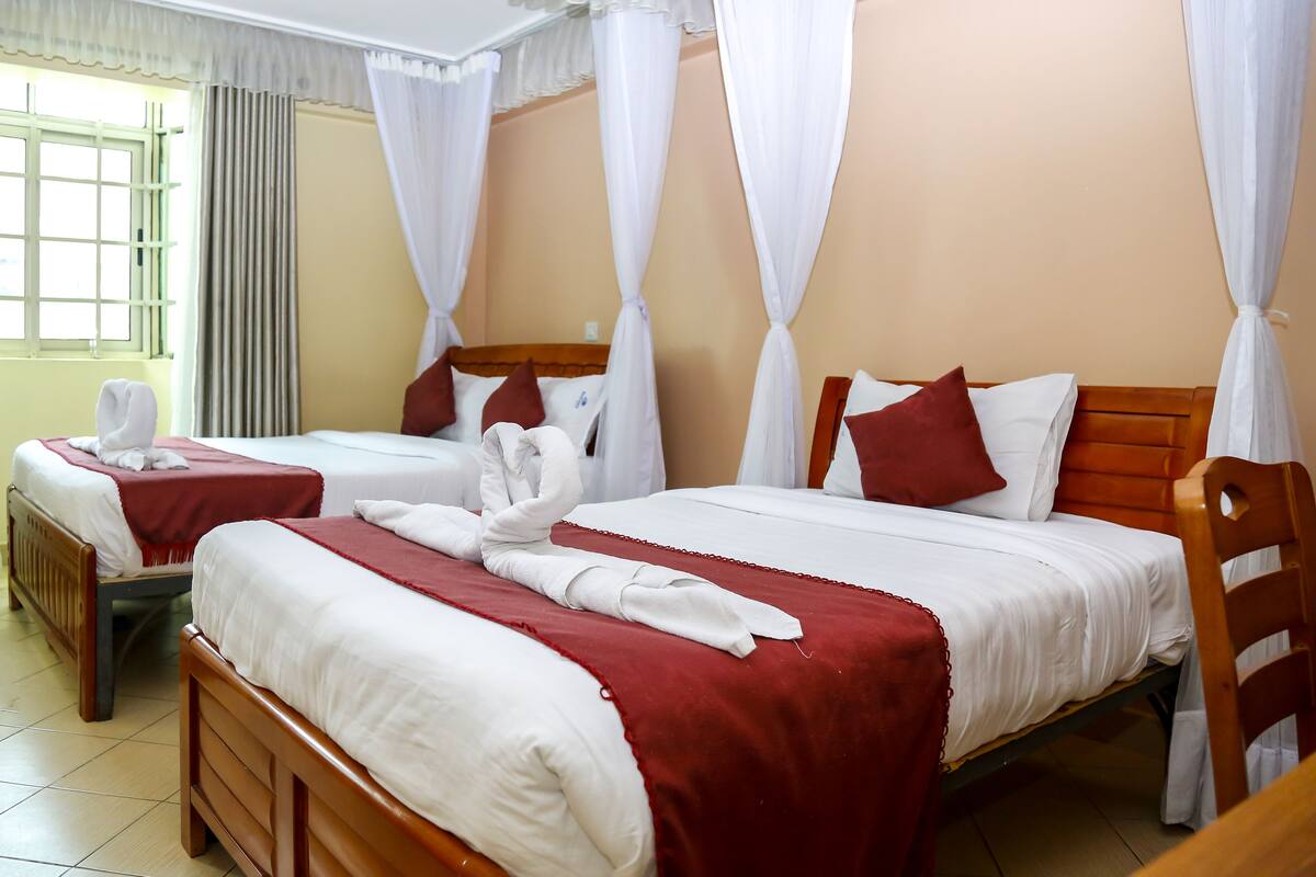 Una habitación de hotel con dos camas, cada una con sábanas blancas y colchas rojas. Hay cortinas blancas y un cabecero de madera.
