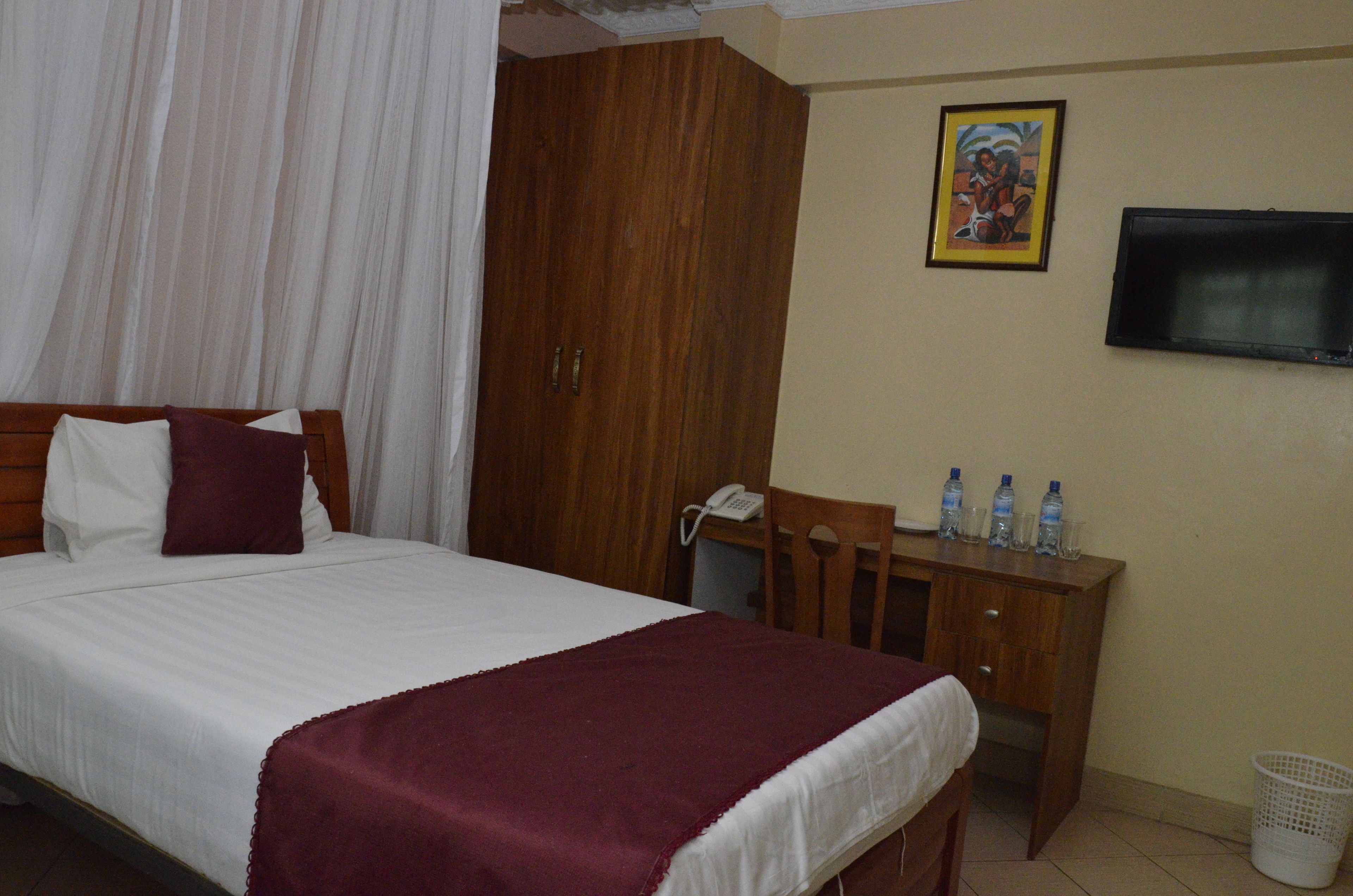 A hotel room with a bed, a wooden wardrobe, a desk with water bottles, a TV, and a wall-mounted painting.