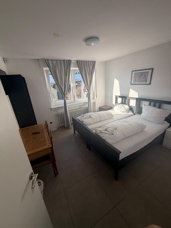 A hotel room with a bed, a desk, a chair, a window with curtains, a radiator, and a framed picture on the wall. - GZ Hostel Bonn (Bonn)