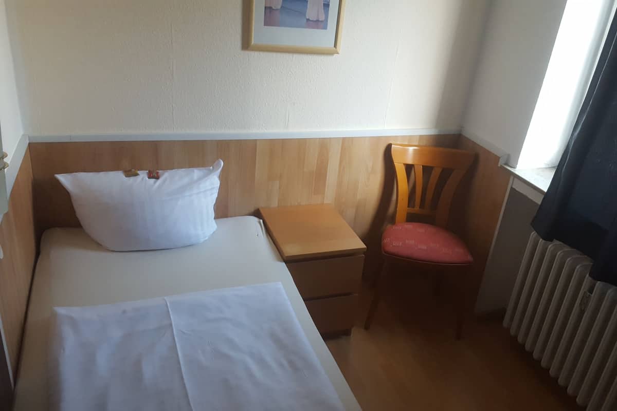 Una habitación pequeña y sencilla con cama, mesita de noche, silla y radiador.