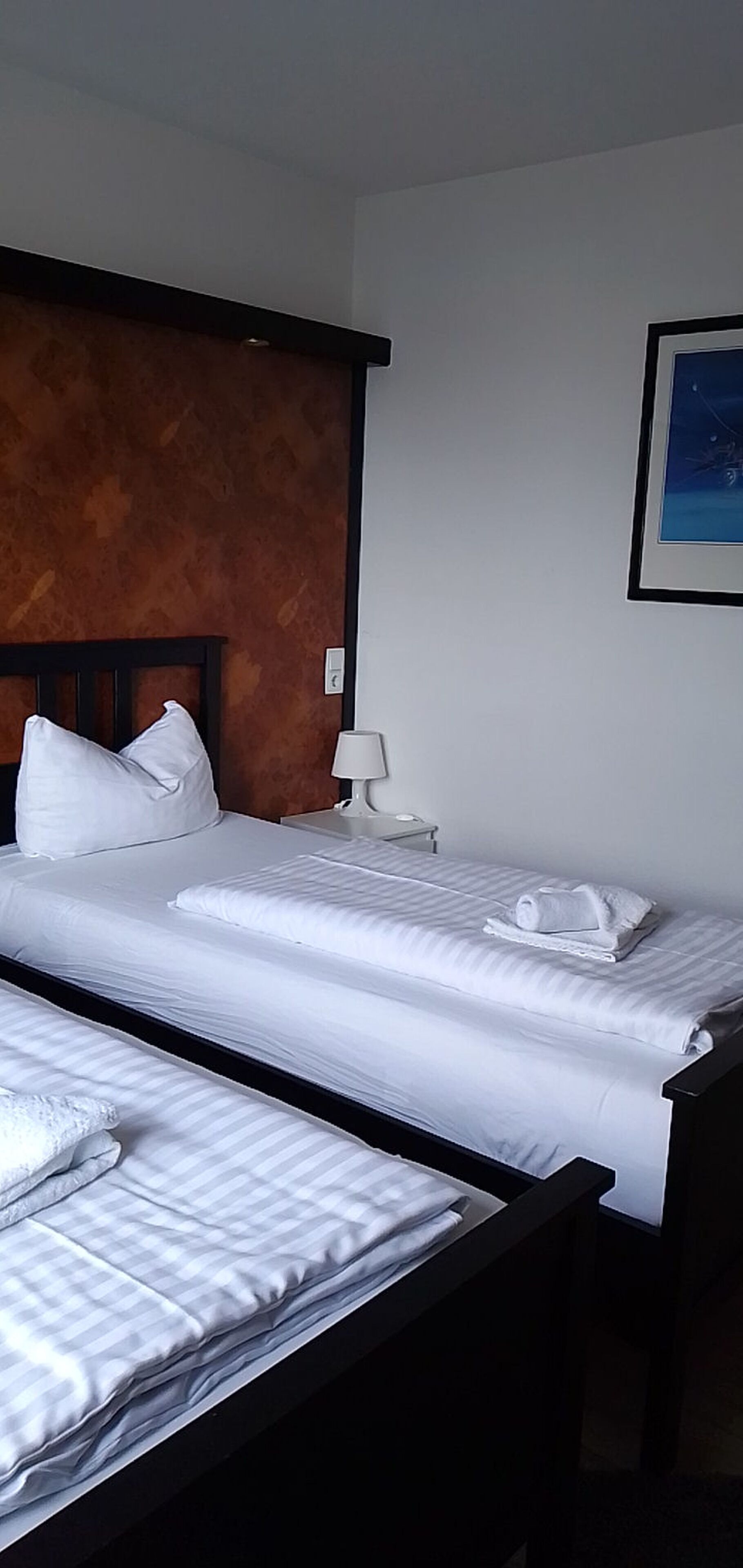 Een hotelkamer met twee bedden, een houten hoofdbord, een nachtkastje, een lamp en een lijst met een foto aan de muur.