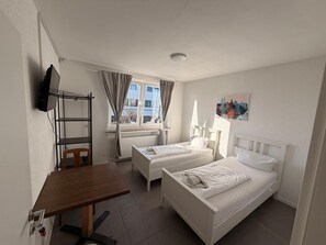 Una habitación pequeña y limpia con dos camas individuales, un escritorio, una silla, un televisor y una ventana con cortinas.