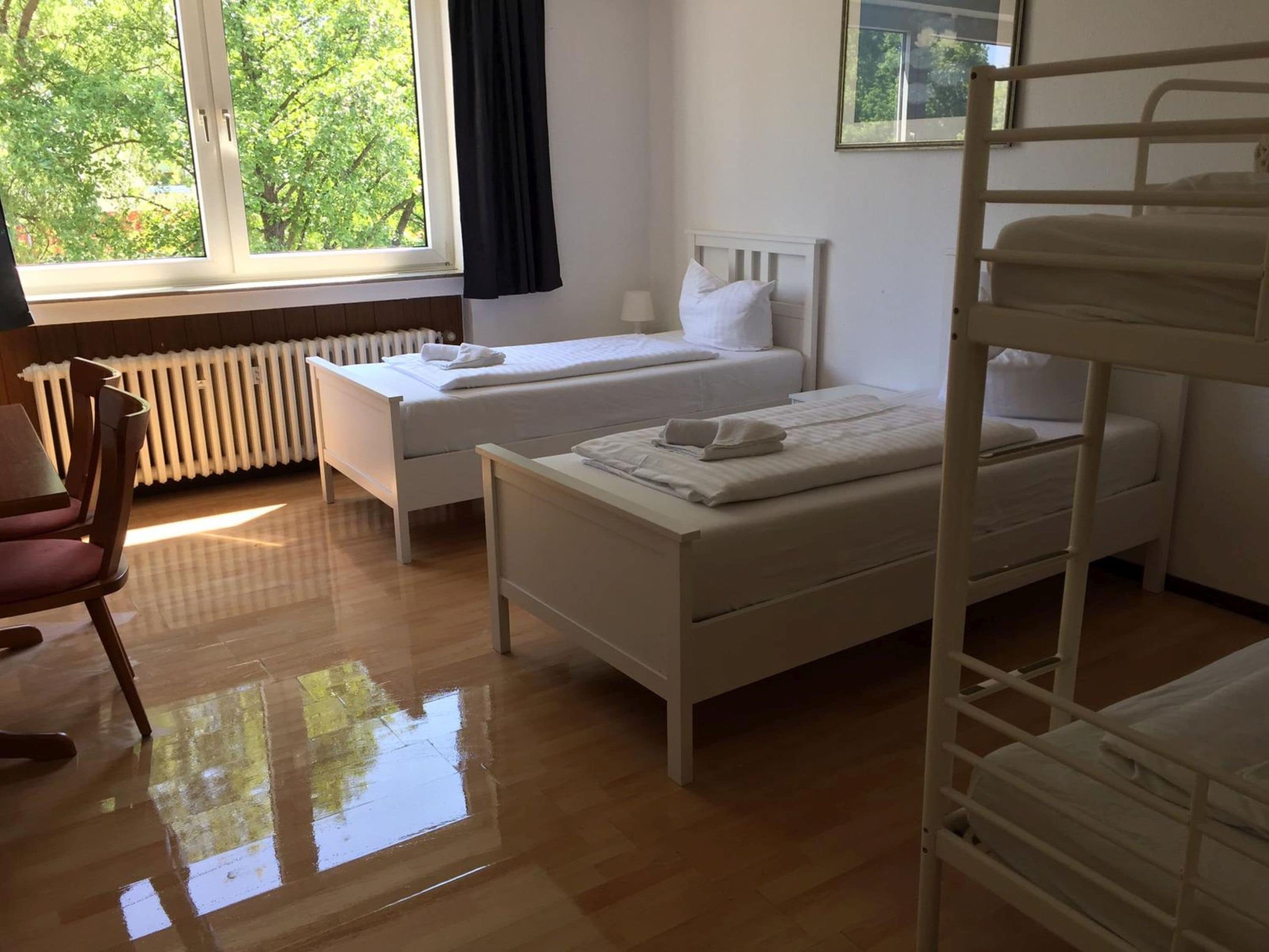 Une chambre d’hôtel avec deux lits simples, une chaise en bois, un radiateur et une fenêtre donnant sur des arbres.