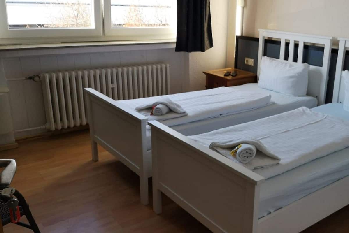 Habitación de hotel con dos camas individuales, suelo de madera, radiador, mesita de noche y ventana con cortinas.