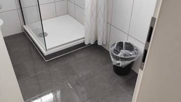 Un baño con ducha de vidrio, paredes de azulejos blancos y un cesto de basura negro.