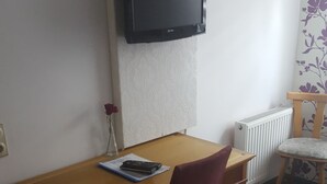 Une pièce avec une table en bois, une chaise rouge, un téléviseur fixé au mur, un radiateur et un papier peint à motifs floraux.