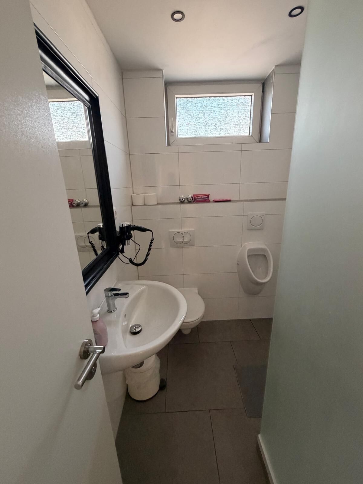 Un baño pequeño con lavamanos pedestal, inodoro, orinal y un ventanal sobre el inodoro.