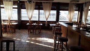 Un restaurant avec des tables et des chaises en bois, de grandes fenêtres avec des rideaux et un sol carrelé.
