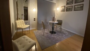 Une pièce avec un plancher en bois, une porte blanche, une petite table ronde sur laquelle se trouve un vase, deux chaises et des tableaux encadrés accrochés au mur.