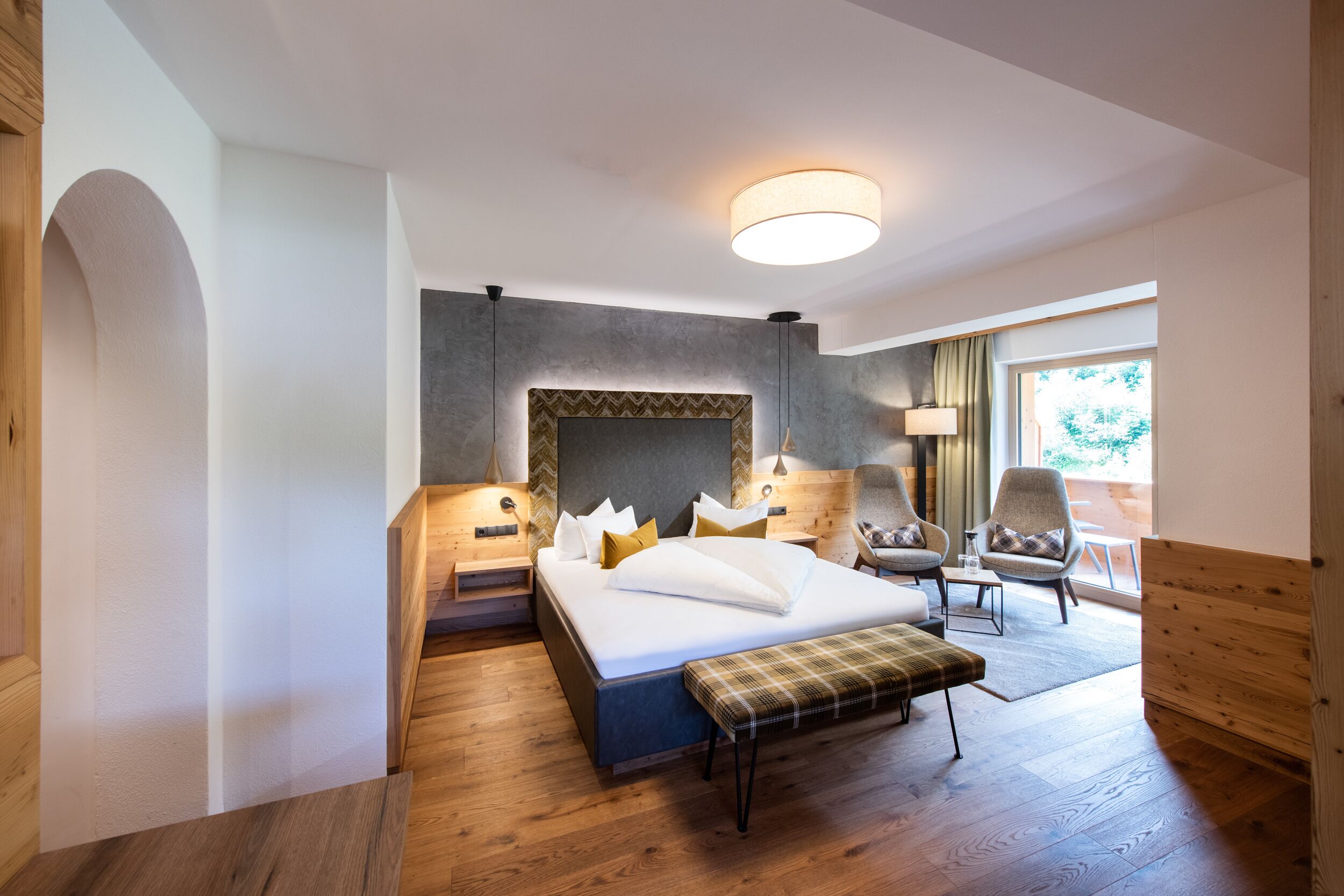Ein modernes Hotelzimmer mit einem großen Bett, einer Bank, einem Sessel und Blick ins Freie.