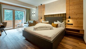 Une chambre moderne dotée d’un grand lit, d’une tête de lit en bois, d’un coin salon avec une chaise et une table, et d’un balcon offrant une vue sur les arbres.