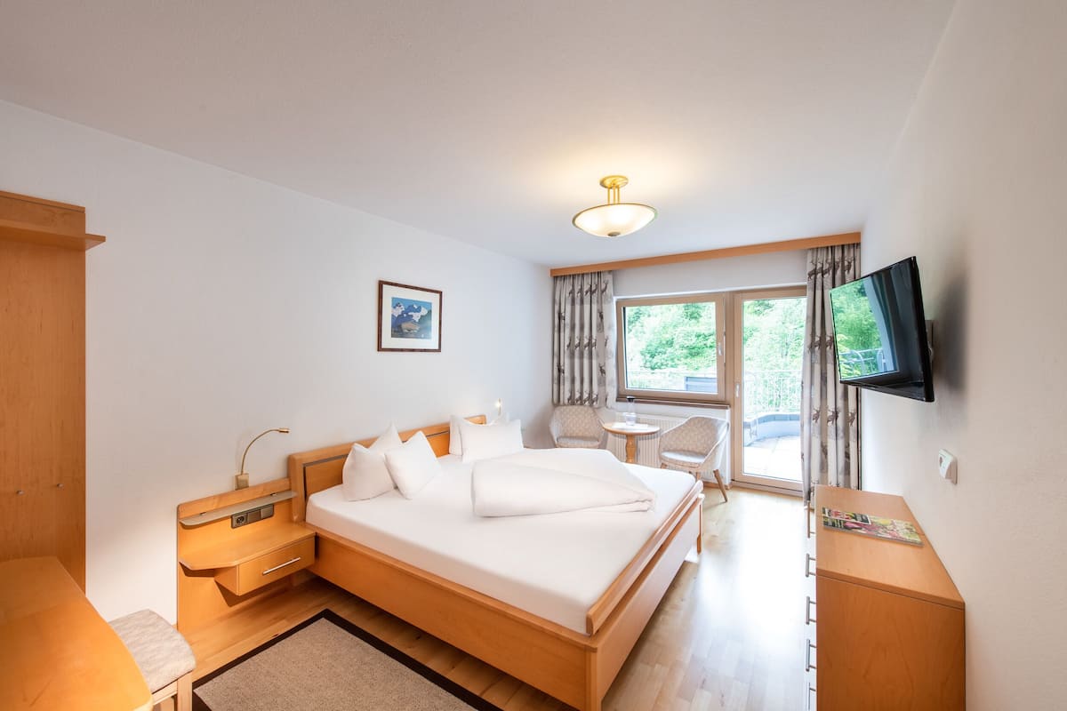 Une chambre d’hôtel avec un lit en bois, une table de chevet, une chaise, une télévision et une fenêtre avec des rideaux.