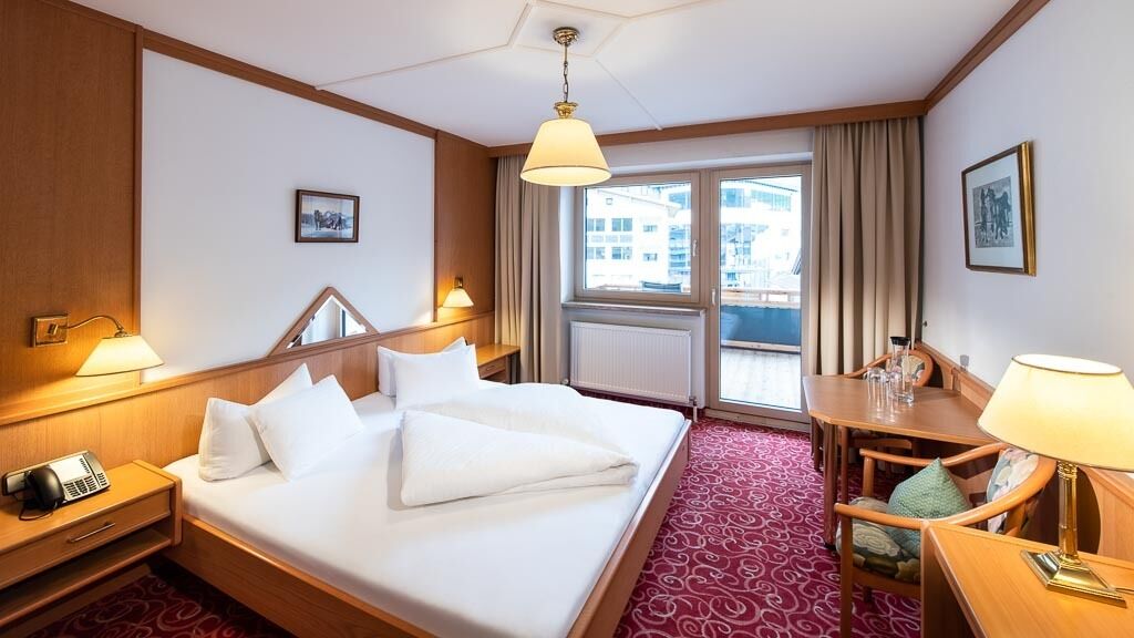 A hotel room with a large bed, a desk with a lamp, a telephone, and a window with curtains.