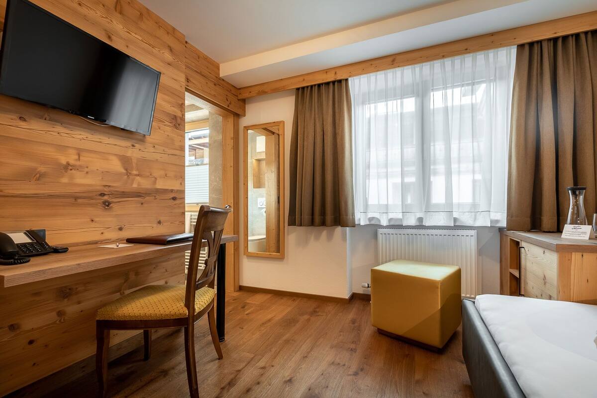 Une chambre d’hôtel avec un bureau en bois, une chaise, un lit, un téléviseur fixé au mur et une fenêtre avec des rideaux.