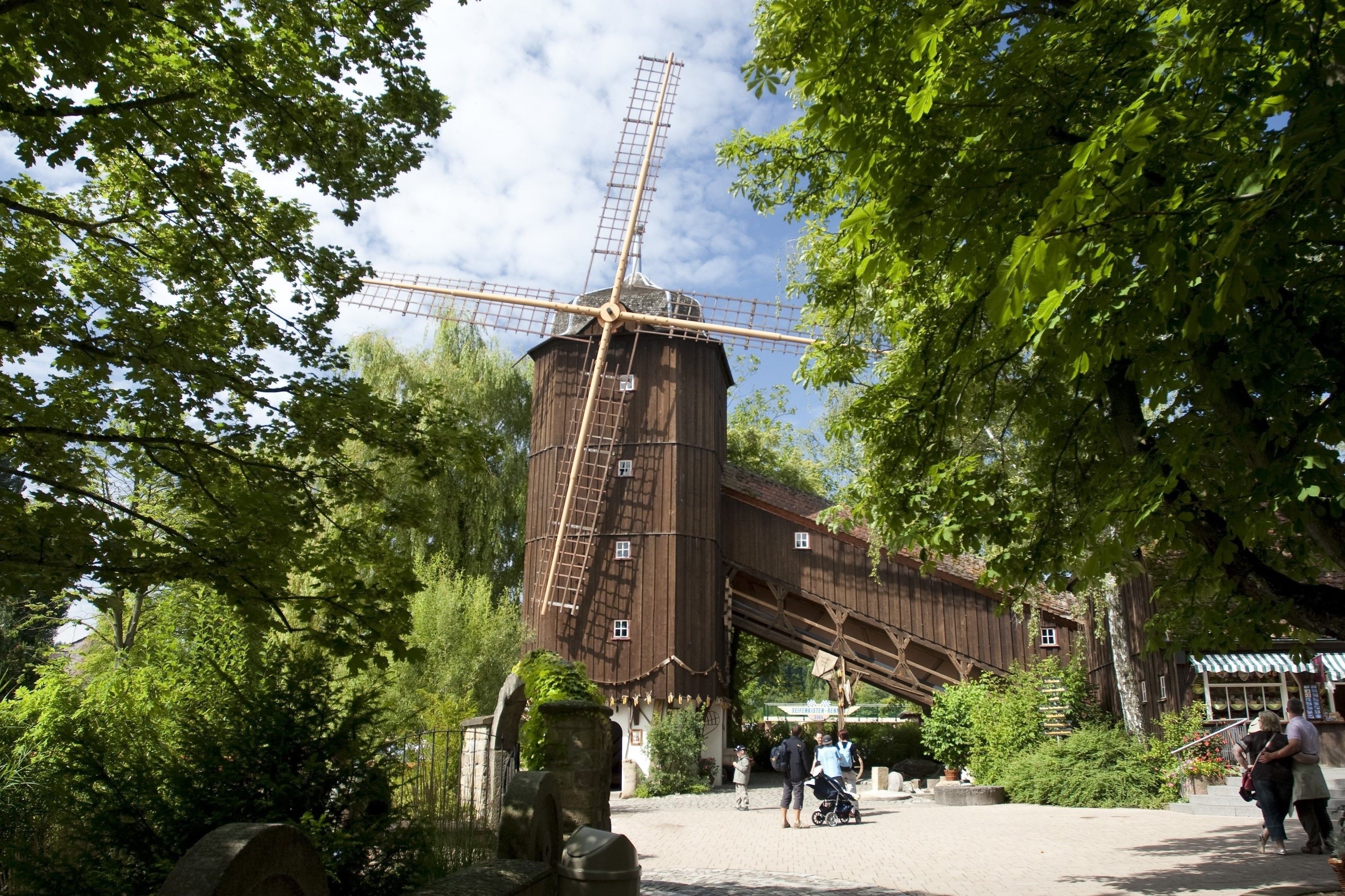 Un moulin à vent traditionnel en bois, doté d’une structure en treillis, entouré d’une végétation luxuriante et d’une zone pavée où se trouvent des personnes et une poussette.