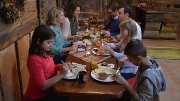 Un grupo de personas cenando en una mesa de madera en un entorno rústico con una pared de ladrillo y bancos de madera.