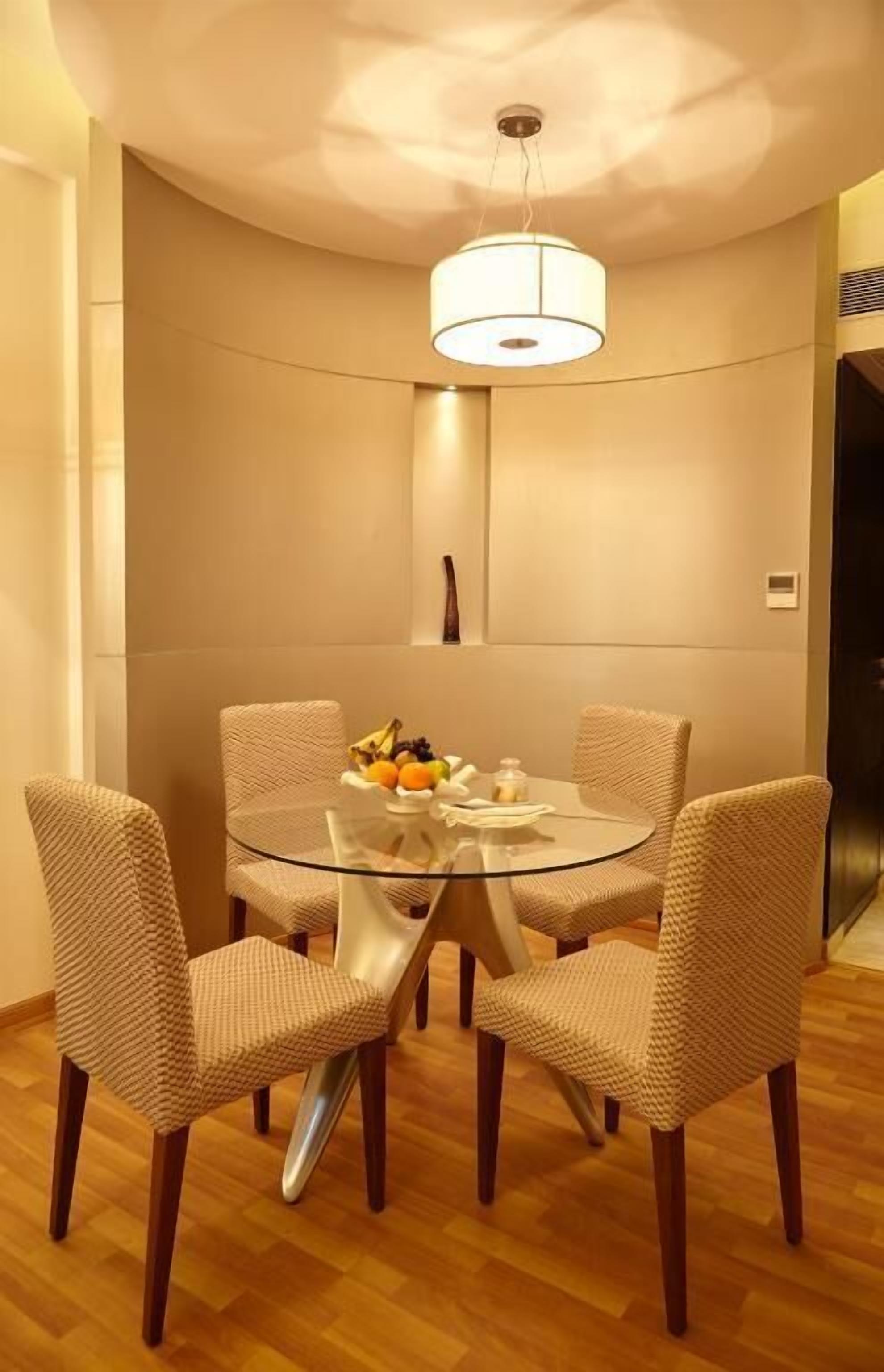 Un coin repas avec une table ronde en verre, entourée de quatre chaises en osier, d’un saladier rempli de fruits et d’un luminaire suspendu.
