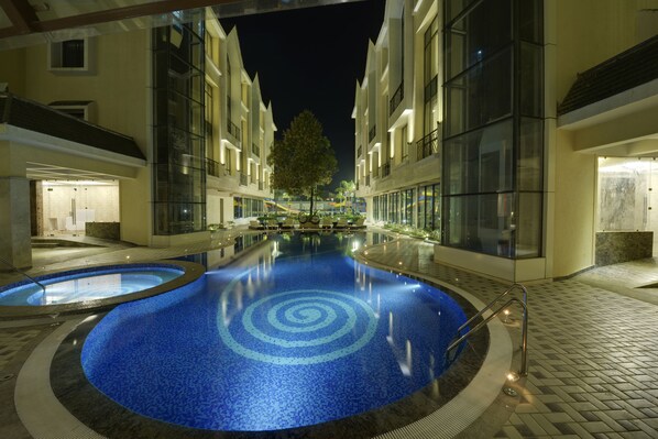 A nighttime swimming pool area with a circular design, surrounded by modern buildings with glass facades and a tree in the center. - Meritas Picaddle Resort - Lonavala (Mawal)