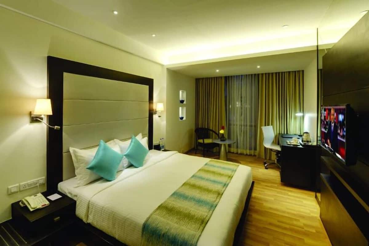 A hotel room with a large bed, bedside tables, a desk with a chair, a TV, and a window with curtains.