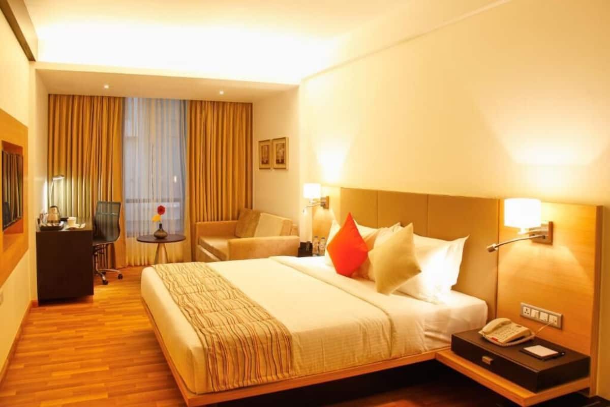 A hotel room with a large bed, bedside lamps, a desk with a telephone, and a view of the city through the window.