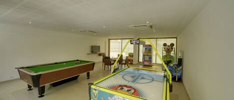 A pool table, a children's arcade, and a seating area in a room with white walls and a tiled floor.