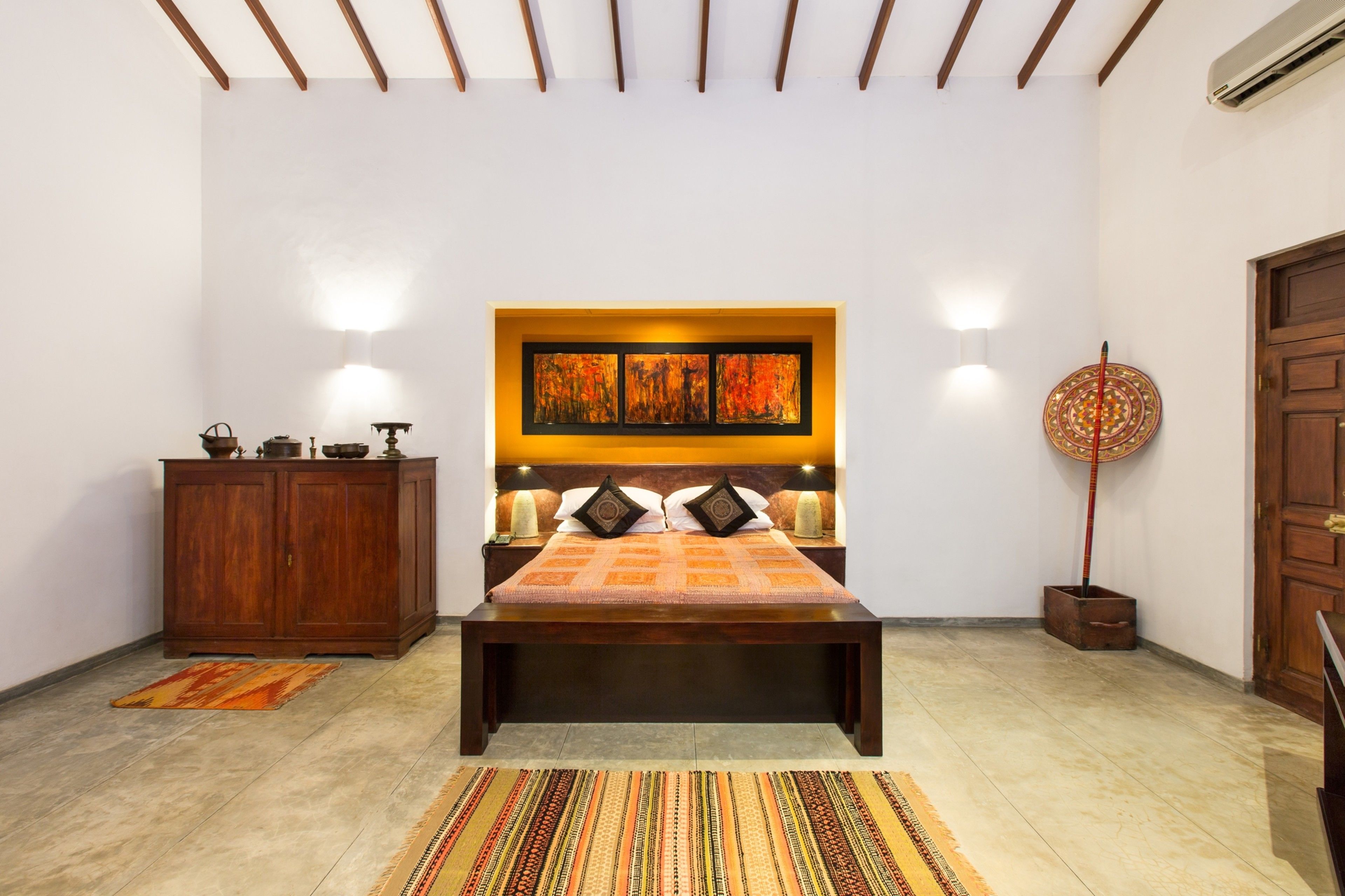 A bedroom with a bed, wooden headboard, bedside lamps, a nightstand, a cabinet, a decorative wall piece, and a patterned rug.