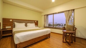 A hotel room with a large bed, wooden headboard, a small table with a chair, and a window with a view of buildings.