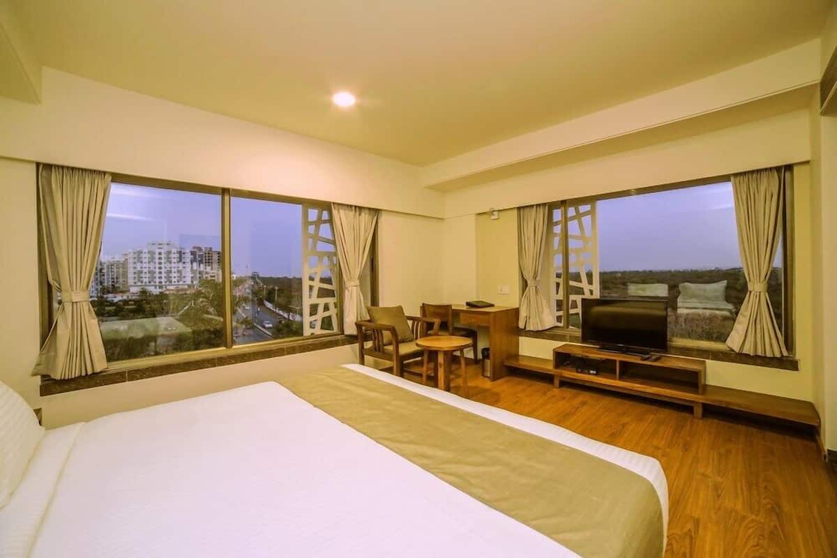 A hotel room with a large bed, a desk, a chair, a TV, and large windows offering a city view.