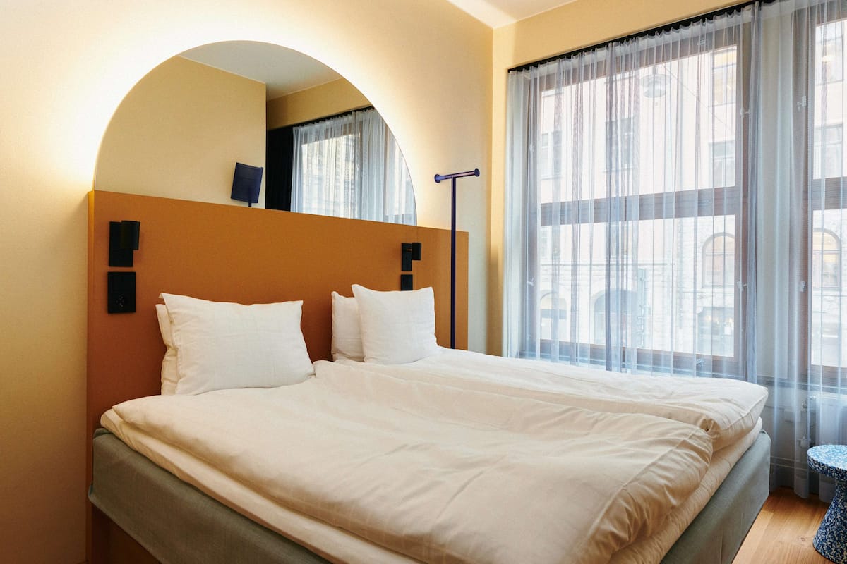 A hotel room with a large bed, a headboard, a bedside table, and a window with sheer curtains.