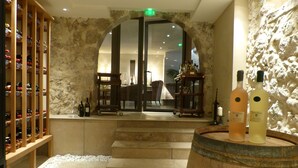 A wine cellar with wooden shelves and a stone wall. There are steps leading to a door, and a wooden barrel in the foreground. - Nouvel Hôtel du Commerce (Castellane)