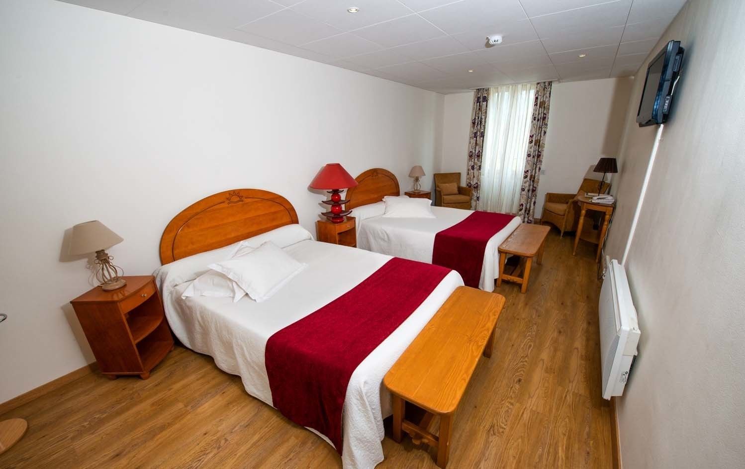 Habitación de hotel con dos camas, cabeceros de madera, mesitas de noche con lámparas, un banco de madera, un televisor montado en la pared y una ventana con cortinas.