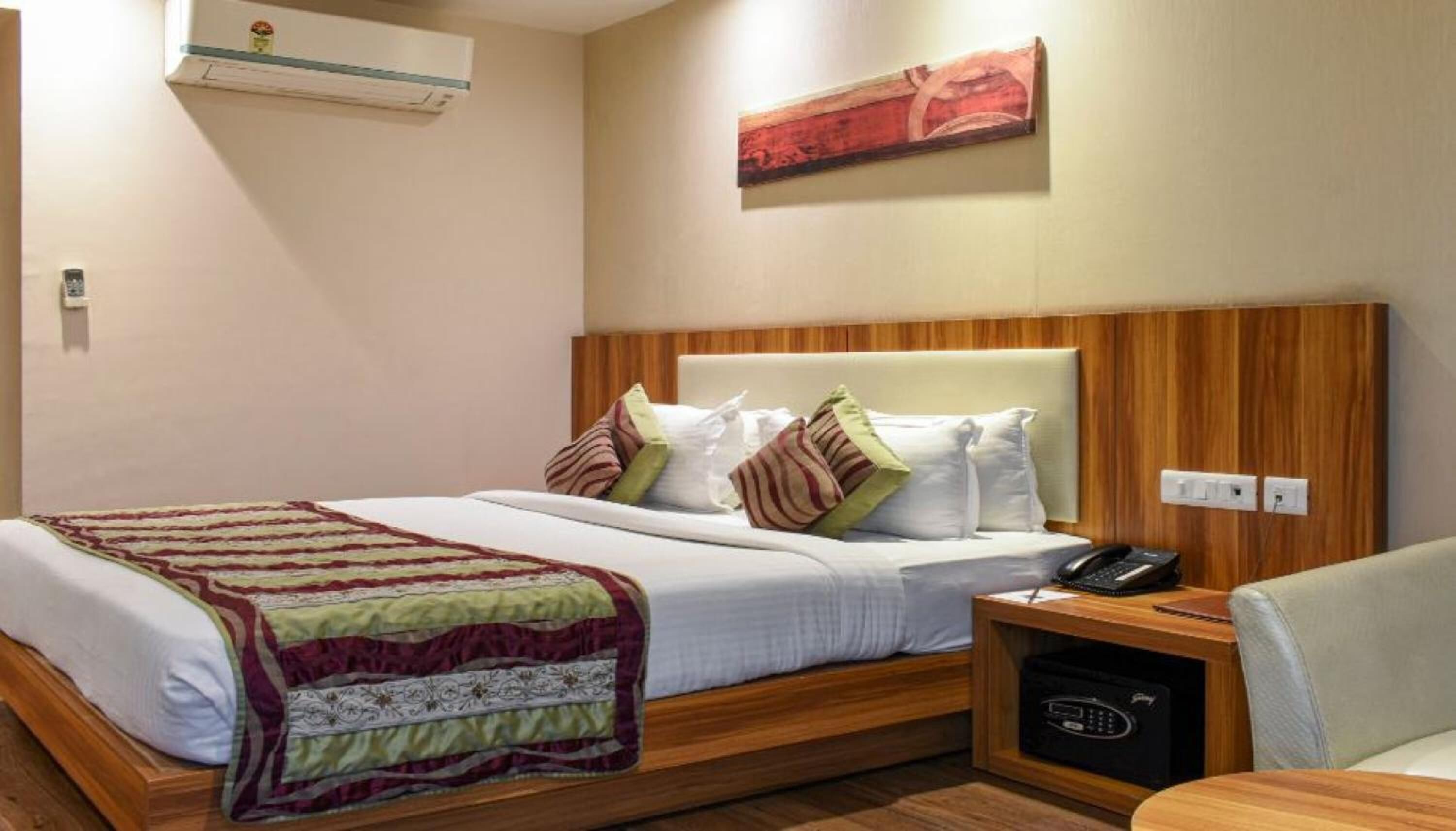 Quarto de hotel com cabeceira de madeira, cama com lençóis brancos, vários travesseiros, mesa de cabeceira com telefone e um pequeno armário.