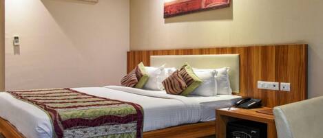 Quarto de hotel com cabeceira de madeira, cama com lençóis brancos, vários travesseiros, mesa de cabeceira com telefone e um pequeno armário.