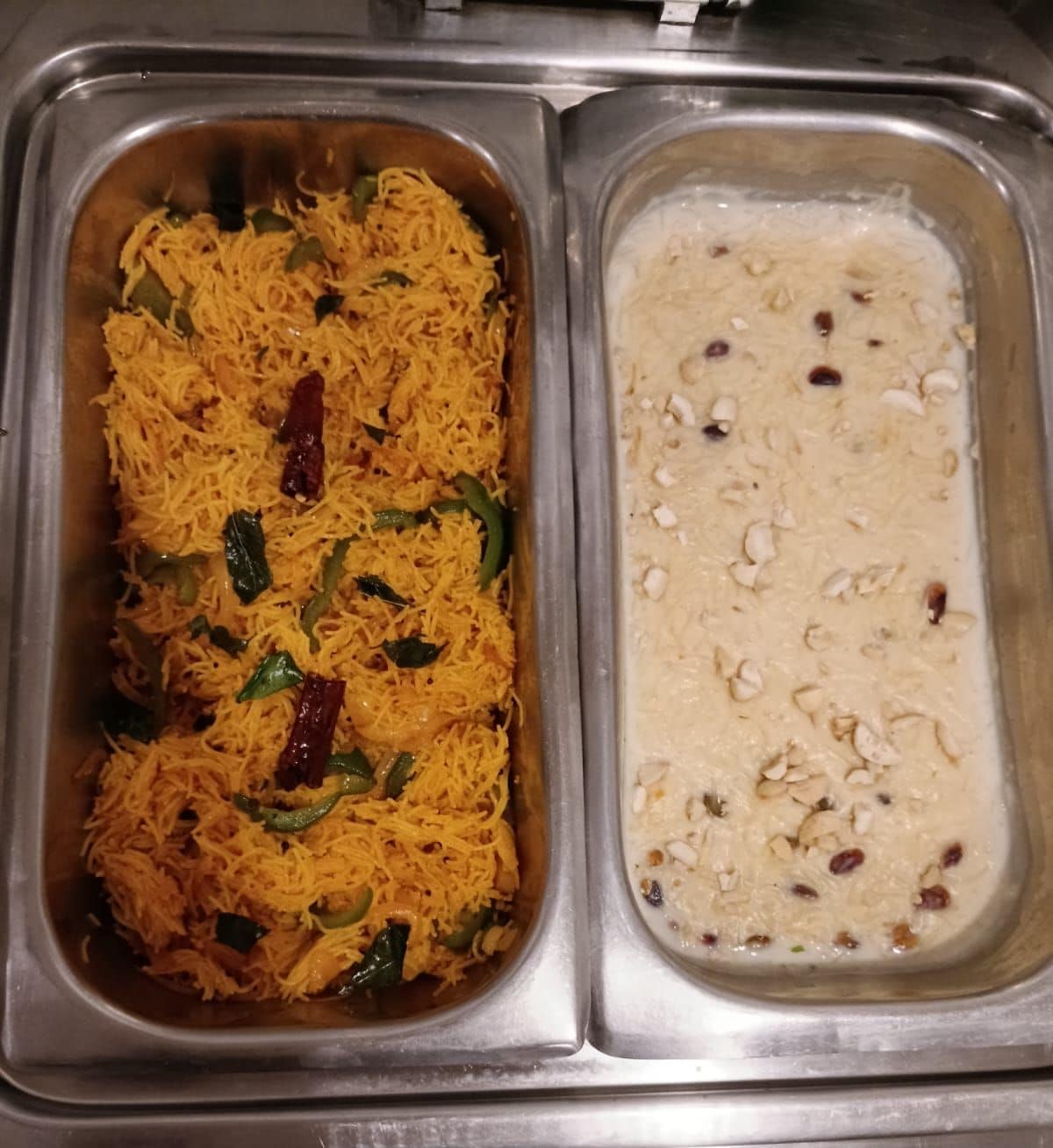Un bufé de acero inoxidable con dos platos: uno parece un arroz amarillo con vegetales verdes y chiles rojos, y el otro es una preparación cremosa con nueces y pasas.