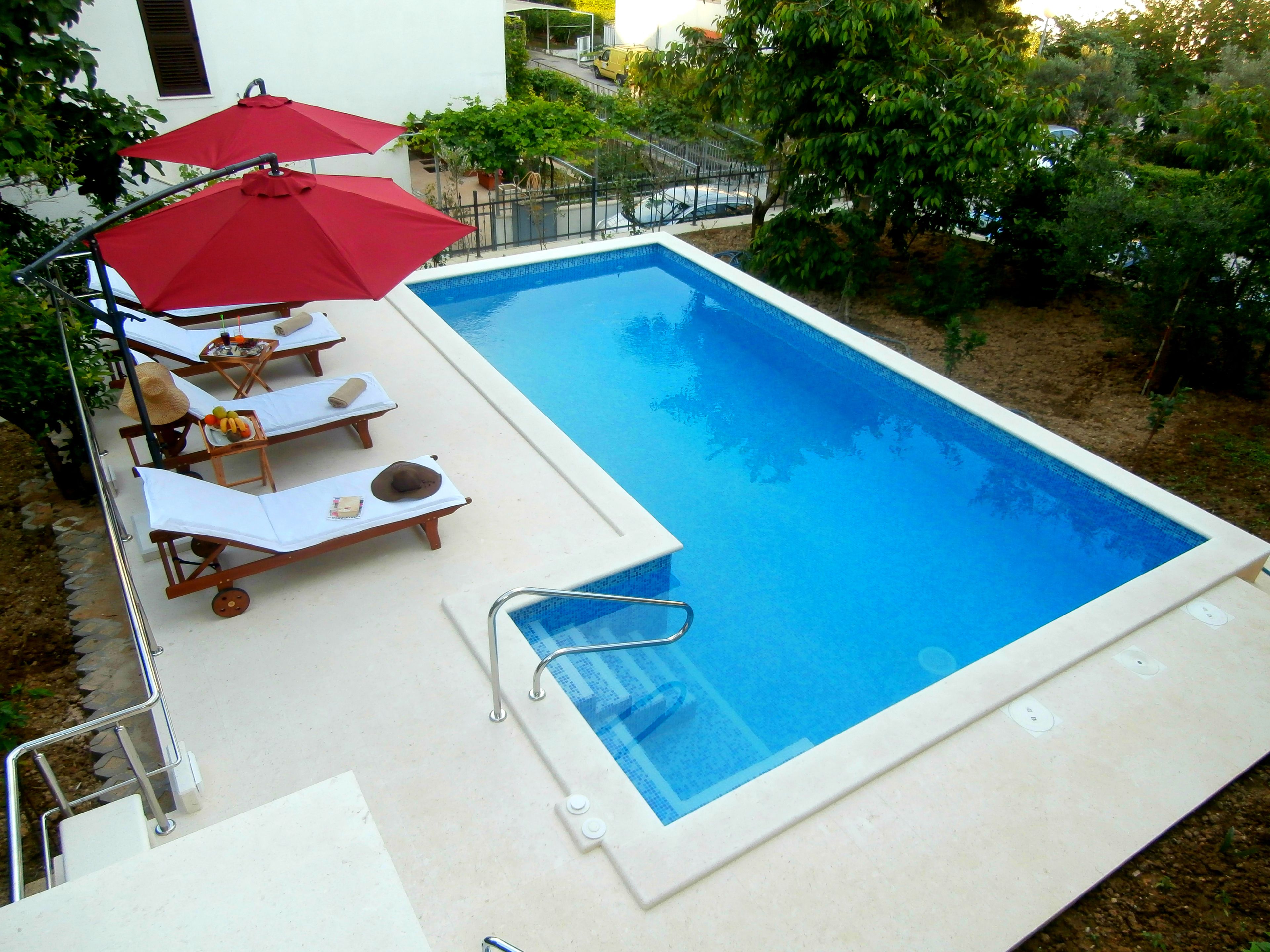 A rectangular swimming pool with a white deck, surrounded by lounge chairs and red umbrellas.