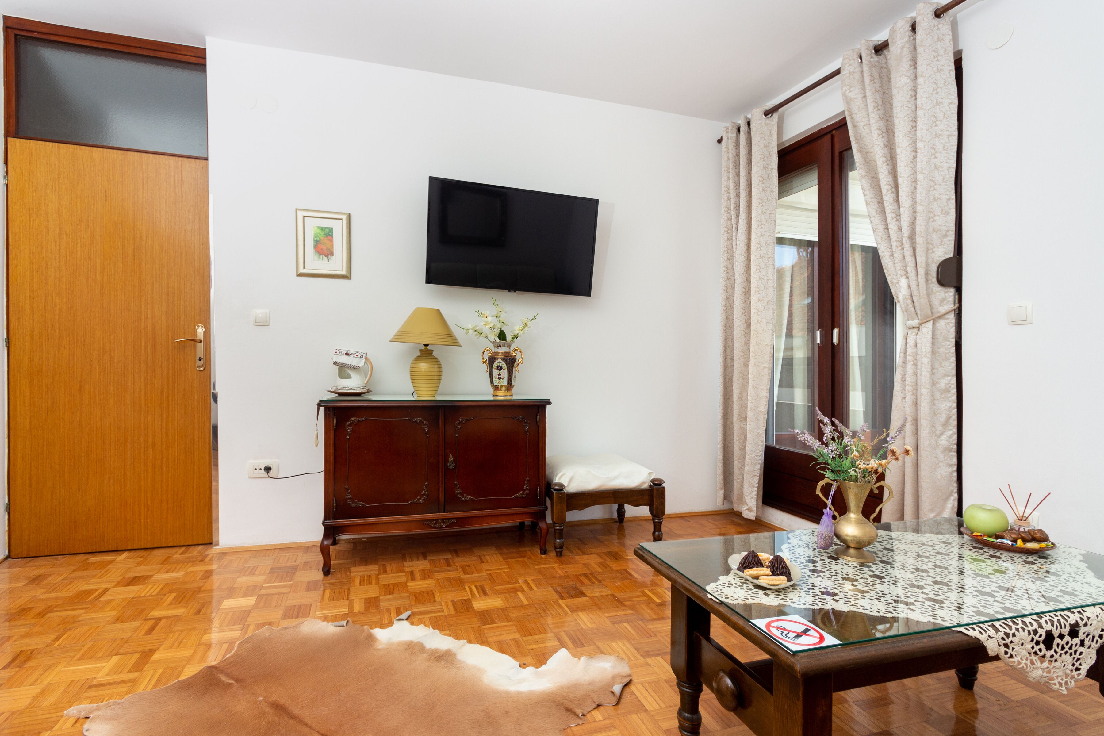 Una habitación con piso de madera, una mesa de vidrio, un sofá, un televisor montado en la pared, un armario y una puerta.