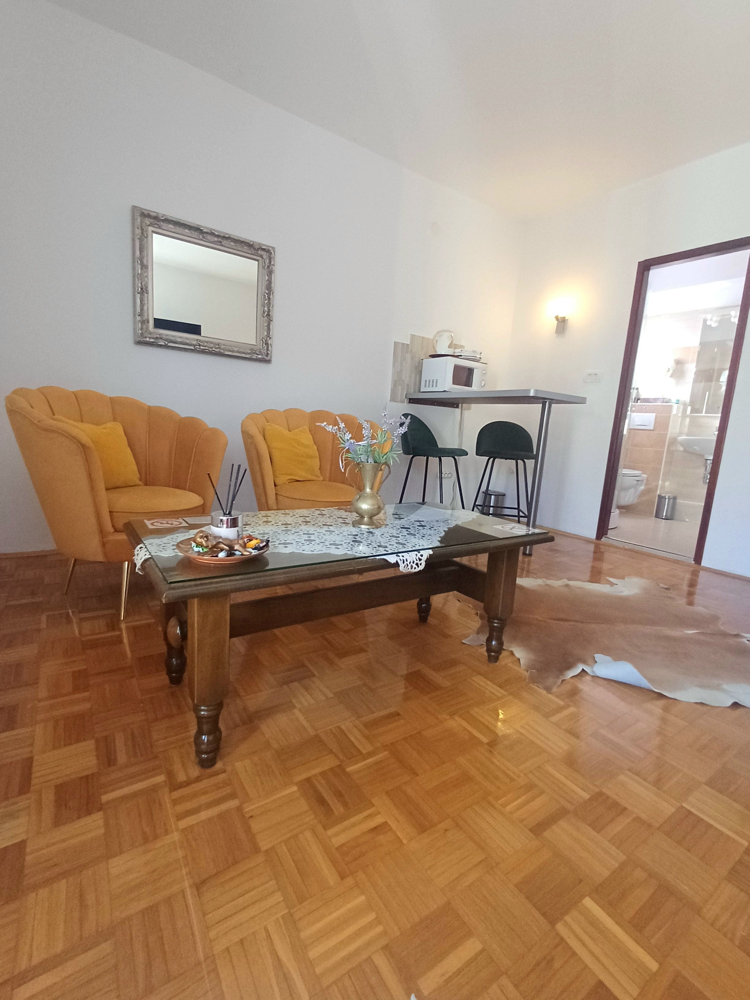 A living room with a wooden herringbone floor, a glass coffee table, two armchairs, a small table with a vase, a microwave, and a framed mirror on the wall.